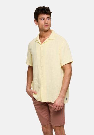 Hombre con camisa de manga corta de color amarillo claro abotonada y pantalones cortos marrones, de pie con una mano en el bolsillo, mirando hacia un lado.