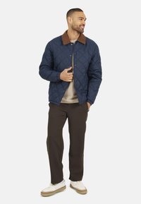 Marine blauwe gewatteerde jas met een bruine corduroy kraag, drukknoopsluitingen en een textuurafwerking, gecombineerd met een bruine broek en witte sneakers.