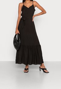 Černé maxi šaty s tenkými ramínky, výstřihem do V, páskem v pase a vrstveným lemem. Kombinované s texturovanou černou kabelkou a černými sandály.