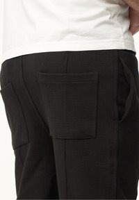 Schwarze Hose mit zwei großen Taschen auf der Rückseite, aus einem weichen, atmungsaktiven Stoff mit matter Textur.