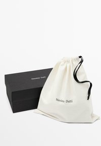 Bomullsdragsäck i off-white med "Massimo Dutti" tryckt i svart, bredvid en svart pappkartong med en slät yta.