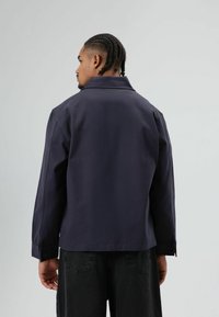 Marineblauwe jas van zachte stof met een kraag, manchetten met knopen en strakke lijnen. Ontworpen met een ontspannen pasvorm en minimale details.