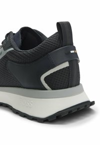 Czarny i szary siateczkowy but sportowy z piętą i pętelką do zakupu, gruba biała podeszwa z wytłoczonym napisem "BOSS" oraz czarna gumowa podeszwa.