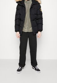 Person in einer schwarzen Daunenjacke mit einem fellbesetzten Kapuzen, grauem Reißverschlussoberteil, schwarzen Hosen, die an den Bündchen aufgerollt sind, und schwarz-weißen Sneakers, steht vor einem einfarbigen Hintergrund.