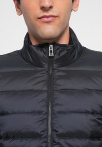Homme portant une veste matelassée noire zippée jusqu'au cou, montrant la moitié inférieure de son visage et de son cou.
