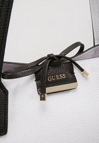 Sac à main texturé noir et blanc avec un détail nœud en cuir noir et une plaque logo "GUESS" dorée.