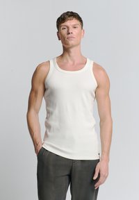 Lichtbeige mouwloze tanktop in een geribbelde textuur, met een ronde halslijn en subtiele stikdetails, gedragen met donkergrijze broek.
