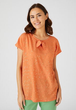 Donna sorridente con indosso una camicetta arancione a maniche corte con stampa quadrettata dorata e un piccolo fiocco frontale, abbinata a pantaloni verdi.