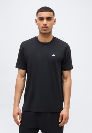 Άνδρας με κοντά μαλλιά που φοράει μια απλή μαύρη μπλούζα με ένα μικρό λευκό λογότυπο στο αριστερό στήθος, στέκεται μπροστά σε ένα απλό φωτεινό φόντο.