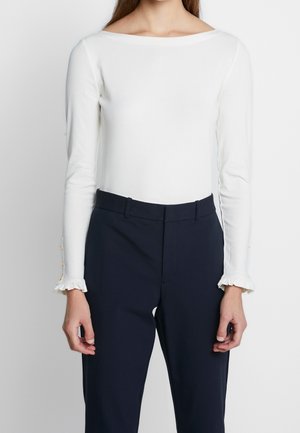 Vrouw draagt een witte top met lange mouwen met geplooide manchetten en gouden knopen, gecombineerd met hoog getailleerde, donkerblauwe op maat gemaakte broek.