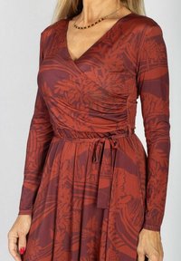 Sukienka kopertowa z długim rękawem w kolorze rdzawego czerwonego materiału, zdobiona botanicznym wzorem, z paskiem taliującym i miękką fakturą. Długość sięga poniżej kolana.