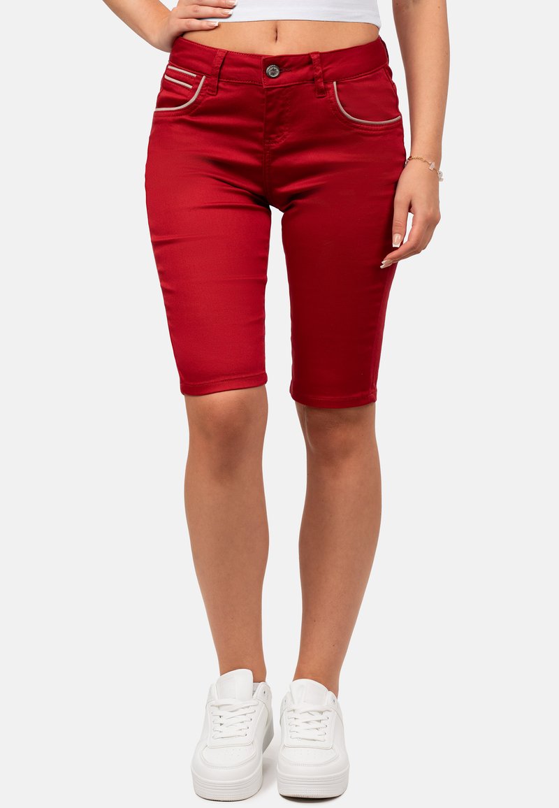 Bermuda shorts in denim rosso con cuciture di accento argento, design aderente e chiusura con un solo bottone anteriore. Indossati con sneakers bianche.