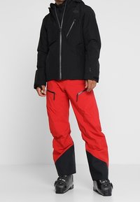 Schwarze wasserdichte Jacke mit Kapuze, Frontreißverschluss und Brusttasche; kombiniert mit roten Hosen mit schwarzen Akzenten und Reißverschlusstaschen, getragen mit Stiefeln.