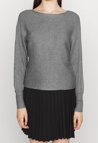 Maglione grigio a coste con maniche lunghe e scollatura ampia, abbinato a una gonna nera plissettata per creare contrasto.