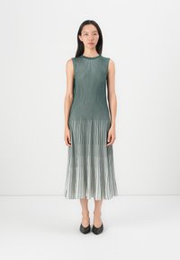 Pleated midi dress in shades of green, featuring vertical stripes, a round neckline, and sleeveless design. Shoes are pointed black heels.