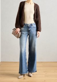 Brauner, flauschiger Cardigan, beiges, figurbetontes Oberteil, hellblaue, weit geschnittene Jeans mit Wäscheeffekt und nuden, spitz zulaufende Absätze, während sie eine kleine graue Tasche hält.