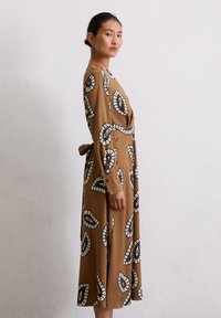 Braunes Midi-Kleid mit weißen und schwarzen kreisförmigen Mustern, langen Ärmeln, seitlichem Bindeband und knöpfbaren Manschetten; glattes Material mit lockerer Passform.