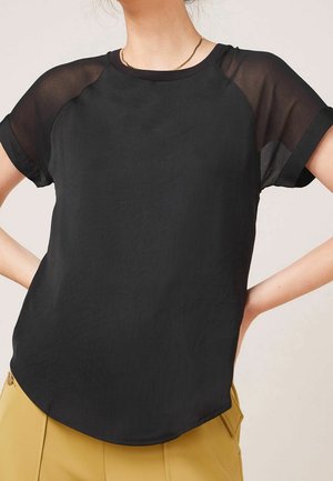 Femme portant un haut noir à manches courtes avec des manches transparentes et un pantalon jaune moutarde, mains reposant sur les hanches, fond clair.