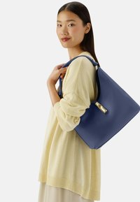 Borsa tote in pelle blu navy con una superficie liscia e dettagli in oro, indossata sopra un top lavorato a maglia giallo chiaro. Design semplice con una forma strutturata.