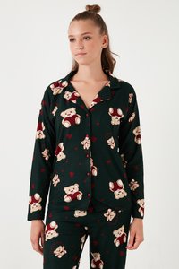 Ung kvinde iført mørkegrønt pyjamasæt med teddy bamse og røde hjerte mønstre, stående mod en ensartet hvid baggrund.