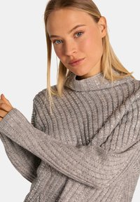 Lichtgrijze geribbelde trui met een hoge halslijn, voorzien van een textuurpatroon en subtiele metallic draden, lange mouwen met brede manchetten.