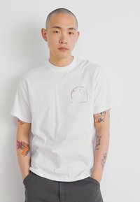 White cotton t-shirt with short sleeves, featuring a small graphic print in the upper left corner. Model wears gray pants and has visible tattoos.