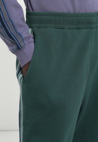 Green cotton sweatpants feature a ribbed waistband, side pockets, and vertical stripes in blue and grey along the side seams.