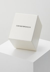 Bílá, texturovaná dárková krabička s mírným leskem. Na přední straně se nachází černé logo "Emporio Armani", které je prezentováno pod úhlem.