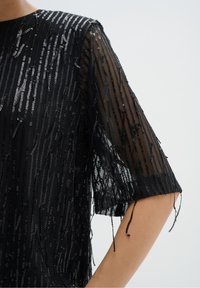 Top trasparente nero con righe di paillettes, maniche corte e dettagli con perline pendenti, che enfatizzano la texture e i design a strati.