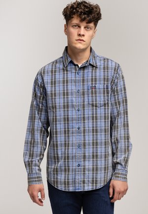 Młody mężczyzna stoi w niebiesko-szarym koszuli w kratę z guzikami i ciemnoniebieskich dżinsach na jednolitym tle.