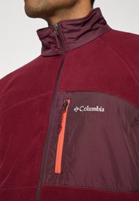 Close-up of a man wearing a maroon Columbia fleece jacket with zippered chest pocket and high collar.