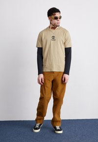 T-shirt di cotone beige con logo Timberland nero, indossato sopra un top a maniche lunghe nero. Pantaloni di corduroy marroni e sneakers nere con suole bianche.