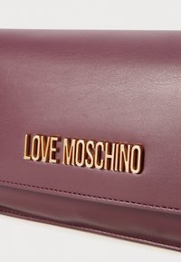 Plommonfärgad läderplånbok med en mjuk textur, prydd med en gyllene "LOVE MOSCHINO"-logotyp på framsidan och sydda detaljer längs kanterna.