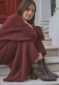 Hnědé kožené kotníkové boty se stříbrným kováním, čtveratou špičkou a blokovým podpatkem. Modelka nosí vínový pletený svetr a sedí na schodech.