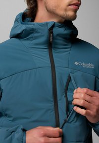 Blaue Columbia Titanium Jacke mit durchgehendem Reißverschluss und Seitentasche. Verfügt über eine Kapuze und ein strukturiertes Gewebe für Wärmeisolierung.
