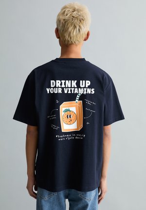 Personne aux cheveux courts et blonds portant une chemise bleu marine avec un dessin animé de boîte de jus d'orange et un texte promouvant les vitamines au dos.