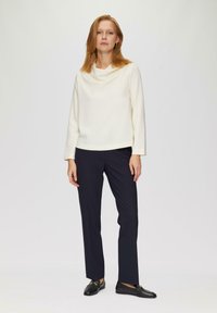 Haut blanc à manches longues avec un col drapé, associé à un pantalon noir à jambes droites et des chaussures noires avec une accent doré. Design simple.