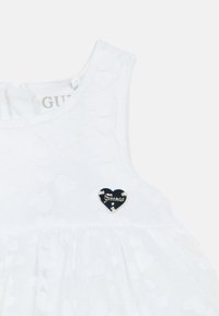 Robe blanche pour enfants avec motif en forme de cœur texturé, un col rond et un badge noir en forme de cœur avec l'inscription "Genuine" en lettres argentées.