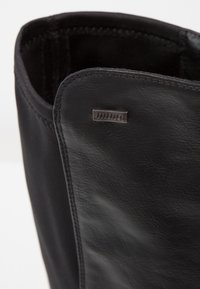 Botte en cuir noir avec une texture lisse, présentant une couture cousue et une petite étiquette logo en métal sur le côté. L'intérieur est doux.