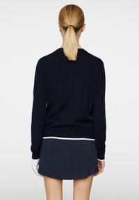 Meripaalto neulepusero, jossa on ribbineuleiset hihansuut ja helma, yhdistettynä meripaalto hameeseen, jossa on valkoinen korostusraita vyötäröllä. Mallin hiukset ovat taakse_muotoiltu.