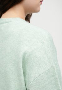 Pull en maille vert clair avec une texture douce, présentant un col rond et des coutures d'épaule abaissées, soulignant une coupe décontractée.