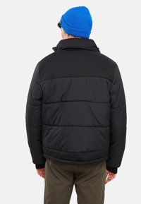 Sort boblejakke med høj krave, quiltet design og ribbede manchetter. Matchet med olivengrønne bukser og en blå strikket hue.