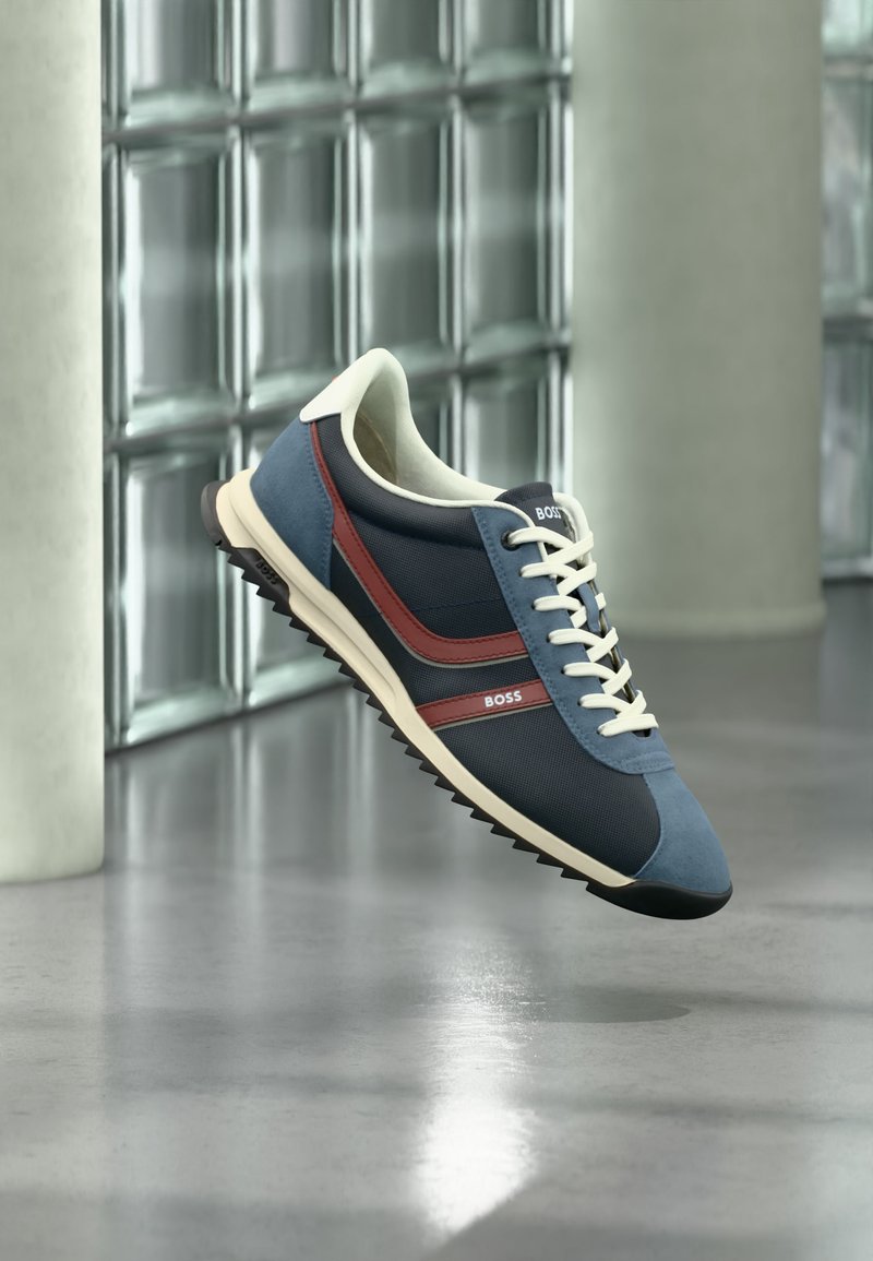 Baskets bleu marine et bleu avec lacets blancs et rayures rouges, de la marque "BOSS", flottant au-dessus d'un sol réfléchissant dans un espace intérieur moderne.
