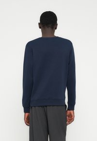 Άνδρας γυρισμένος προς τα πίσω φορώντας μπλε σκουρόχρωμο φούτερ με μακριά μανίκια και σκούρο γκρι παντελόνι, μπροστά σε απλό λευκό φόντο.