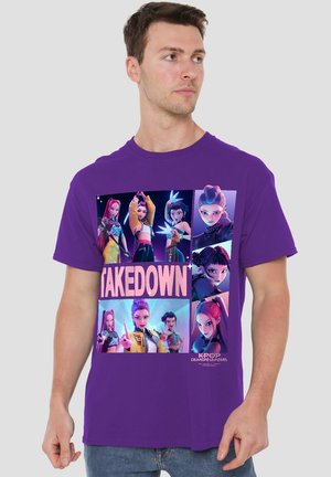 Mann trägt ein lila T-Shirt mit animierten K-Pop-Charakteren in verschiedenen Posen und dem Text „TAKEDOWN“ und „KPOP DEMON HUNTERS“.
