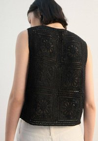Top sin mangas de crochet negro con patrones cuadrados florales texturizados, diseño de tejido abierto y corte corto.