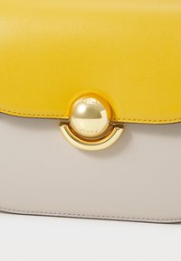 Läderhandväska med ett gult överlock och grå kropp. Har ett guldcirkellås med "FURLA" ingraverat, vilket ger en elegant design och texturkontrast.