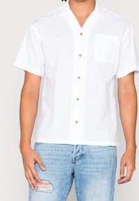 Camicia bianca a maniche corte realizzata in tessuto leggero, con una tasca, cinque bottoni marroni e una vestibilità comoda. Indossata con jeans blu strappati.