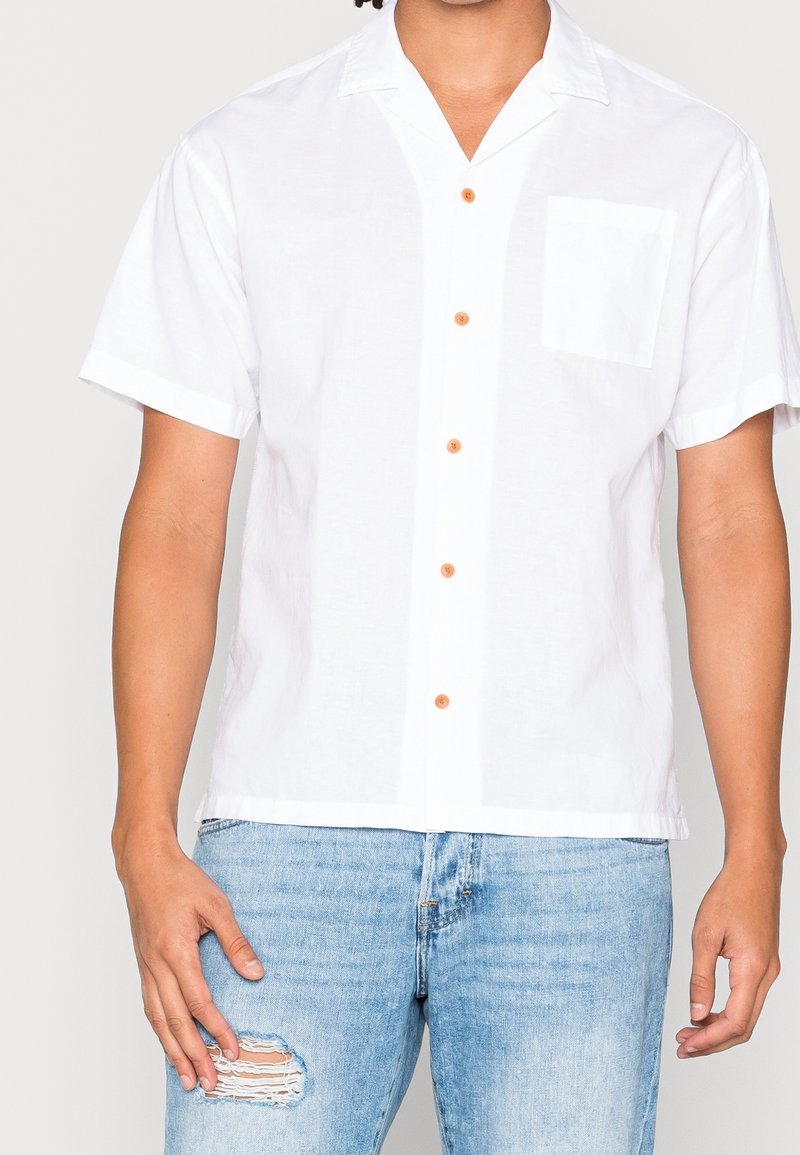 Camicia bianca a maniche corte realizzata in tessuto leggero, con una tasca, cinque bottoni marroni e una vestibilità comoda. Indossata con jeans blu strappati.