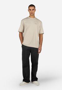 T-shirt bege de manga curta com decote redondo e detalhe de logotipo discreto, combinado com calças largas pretas e ténis brancos.